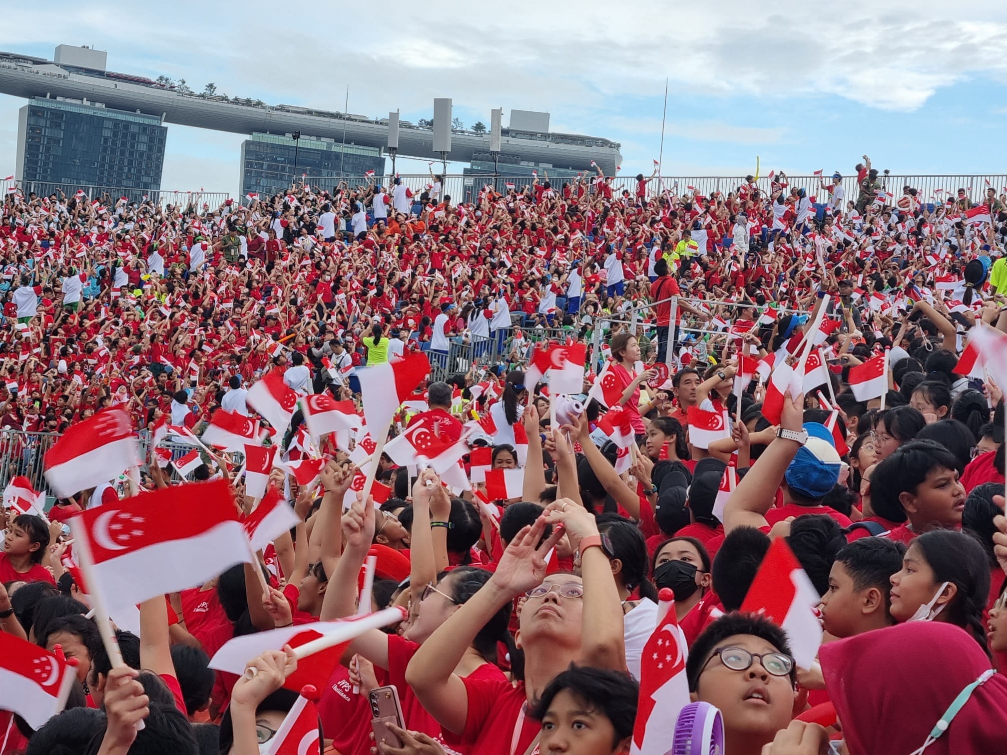 Large crowd in red and white clothing waving Singapore flags at an outdoor celebration event.